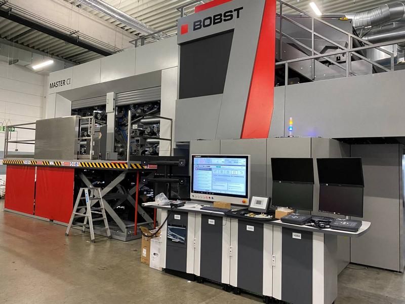 Große BOBST-Industriemaschine in einer Fabrik, davor ein Bedienpult und Computermonitore.