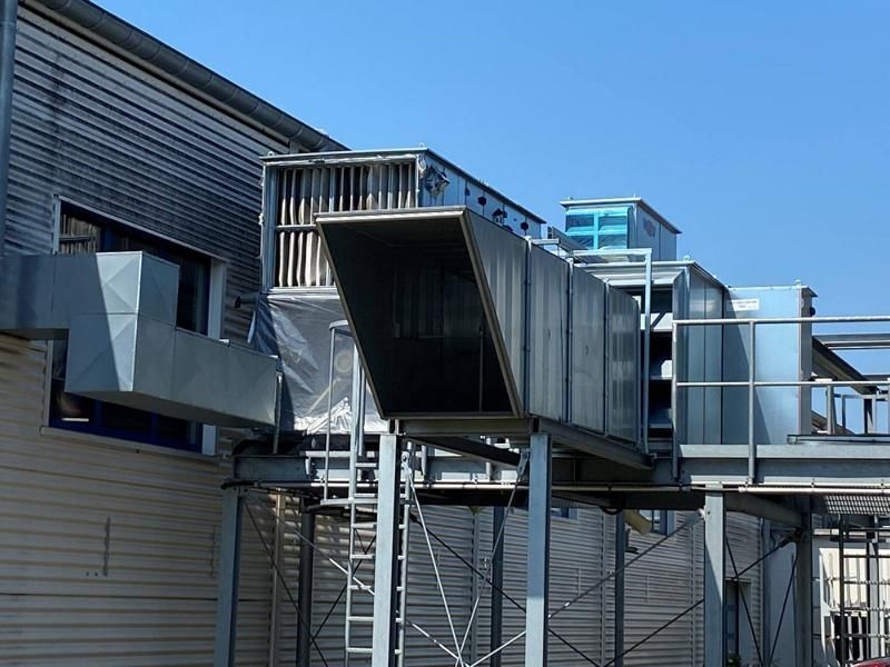 Außenansicht eines Industriegebäudes mit Metallrohren und erhöhtem Laufsteg vor blauem Himmel