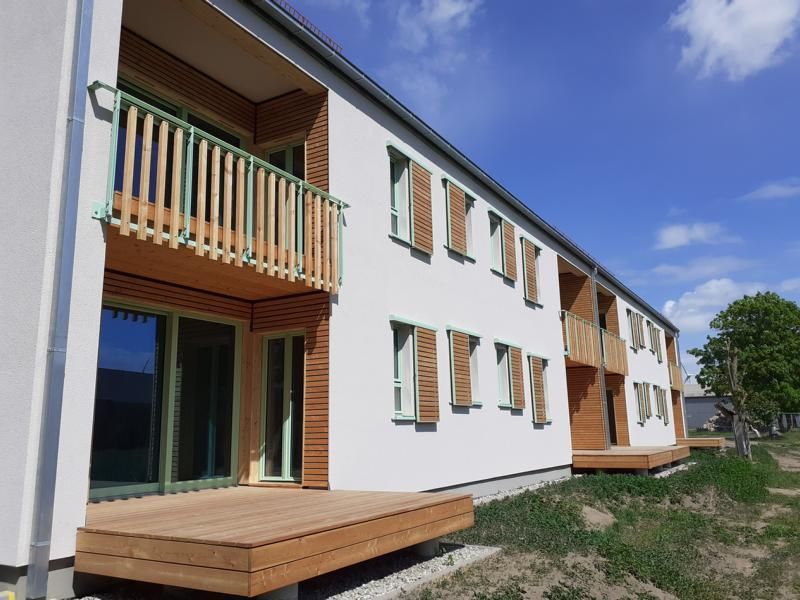 Modernes Mehrfamilienhaus mit weißer Fassade, Holzbalkonen und einer kleinen Terrasse unter blauem Himmel