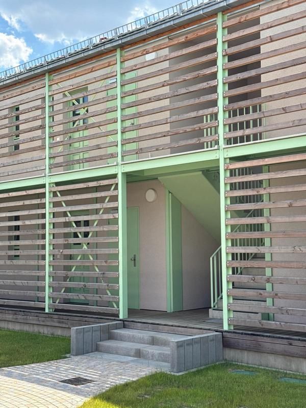 Moderner Gebäudeeingang mit grüner Lamellenfassade und kleinen Stufen, die zu einem zurückgesetzten Eingang führen