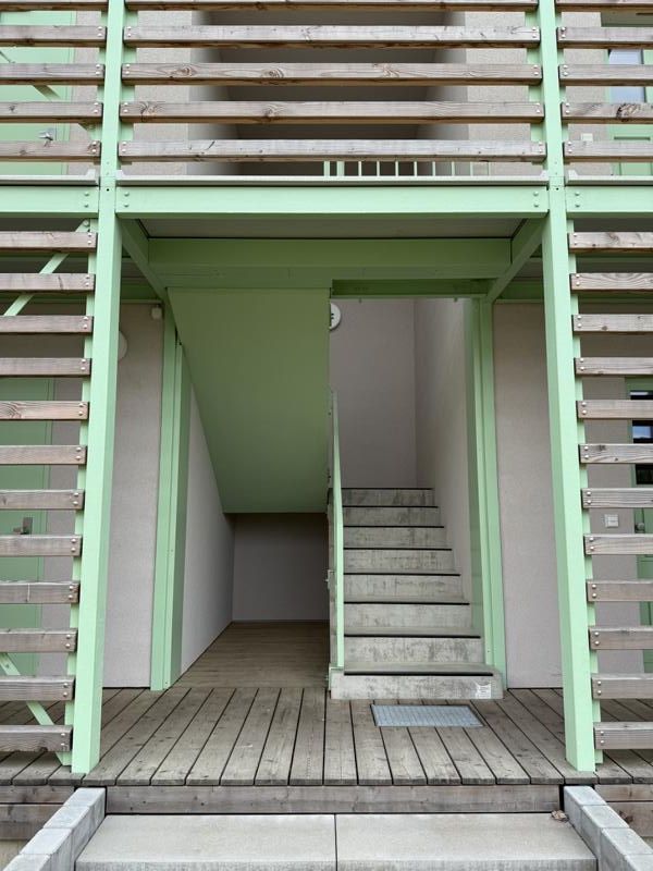 Grüne Holztreppe und Podest unterhalb einer Terrasse, mit zentralem Eingang und seitlichen Stufen.