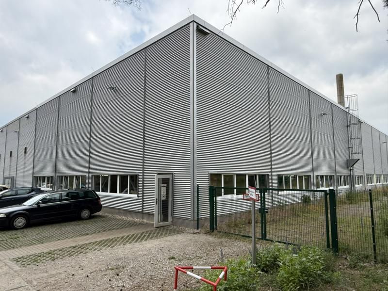 Graues Industriegebäude mit schrägem Dach, geparktem Auto und umzäuntem Eingang unter bedecktem Himmel