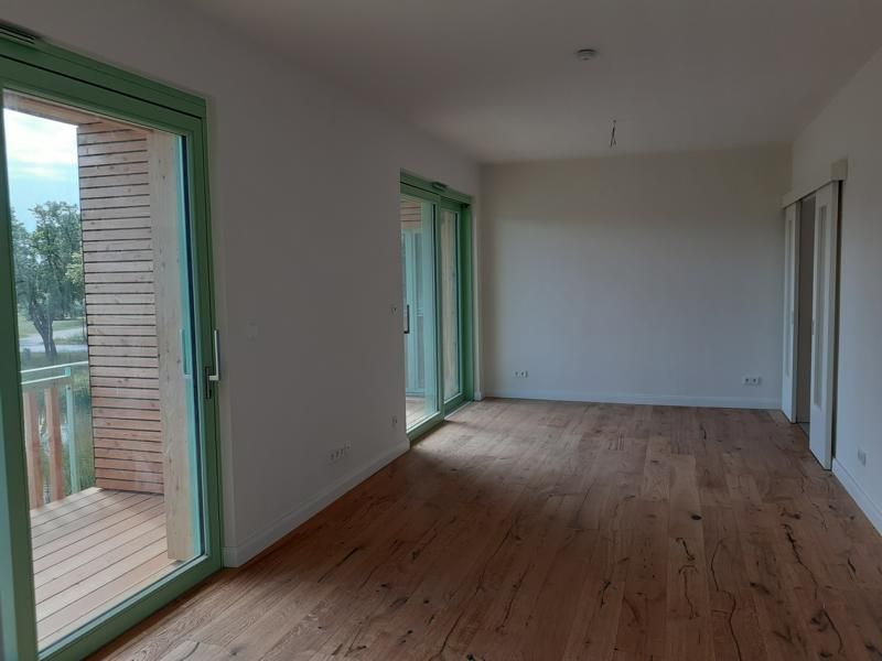 Leeres, modernes Zimmer mit Holzboden, weißen Wänden und grün gerahmten Glasschiebetüren, die auf einen Balkon führen.