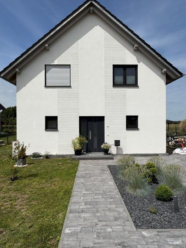 Modernes weißes Haus mit dunklem Dach, mittig angeordneter Eingangstür und einem Steinweg durch einen kleinen Garten.