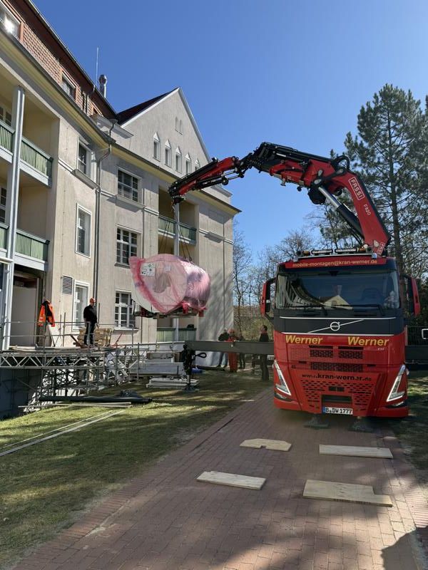 Ein roter Kranwagen hebt einen rosa verpackten Gegenstand neben einem Wohnhaus an einer sonnigen Straße an.