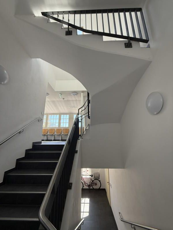 Treppenhaus mit schwarzen Stufen und weißen Wänden, Blick nach oben zu einem Treppenabsatz mit Geländer und Fenster.