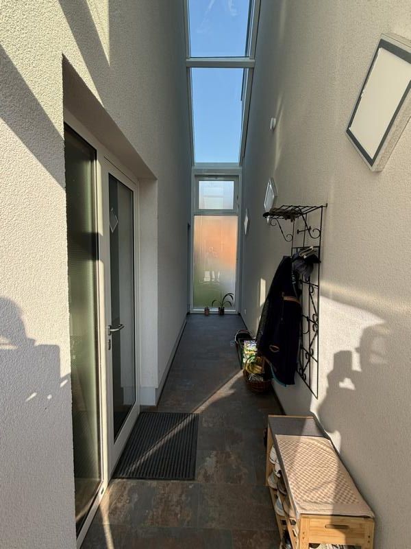 Schmaler Außengang mit Glasoberlicht, Garderobe, Bank und Sonnenlicht, das Schatten auf beige Wände wirft.