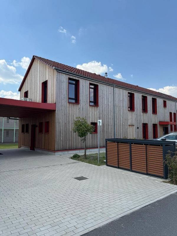 Modernes Holzgebäude mit rot umrandeten Fenstern und einem gepflasterten Innenhof unter blauem Himmel.