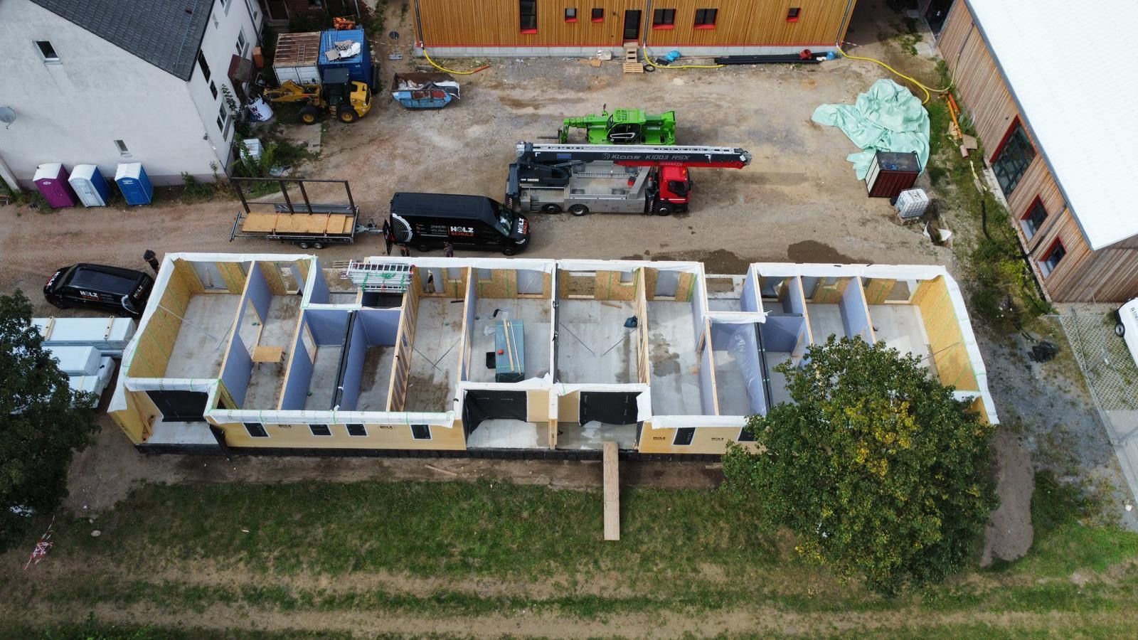 Baustelle aus der Luft: unfertiges, gelb umrandetes Gebäude, Kran, Fahrzeuge und Baumaterialien.