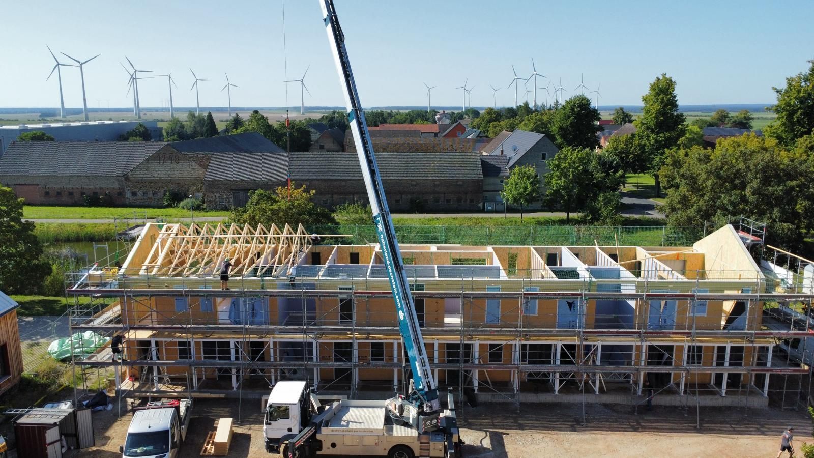 Der Bau eines Holzrahmengebäudes mit einem Kran. Im Hintergrund sind Windkraftanlagen und ein Dorf zu sehen.
