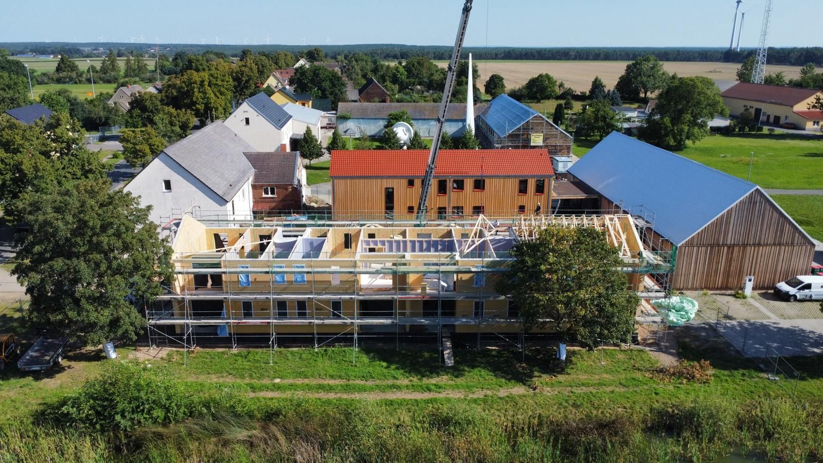 Baustelle mit einem teilweise errichteten Holzgebäude, Kran und ländlicher Umgebung.