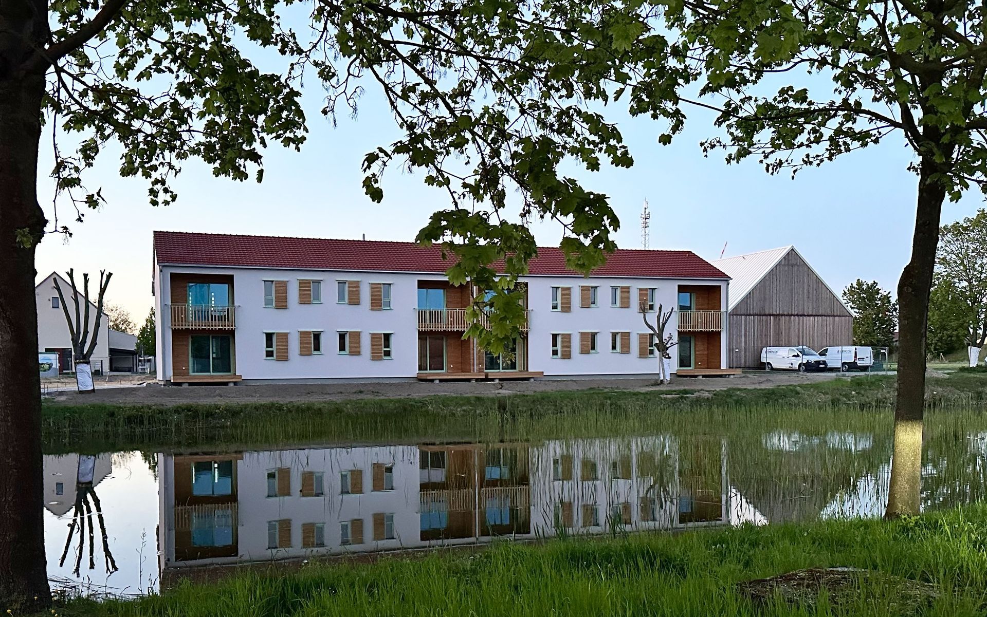 Moderne weiße Gebäude mit roten Dächern spiegeln sich im ruhigen Wasser, im Hintergrund ist eine Scheune zu sehen.