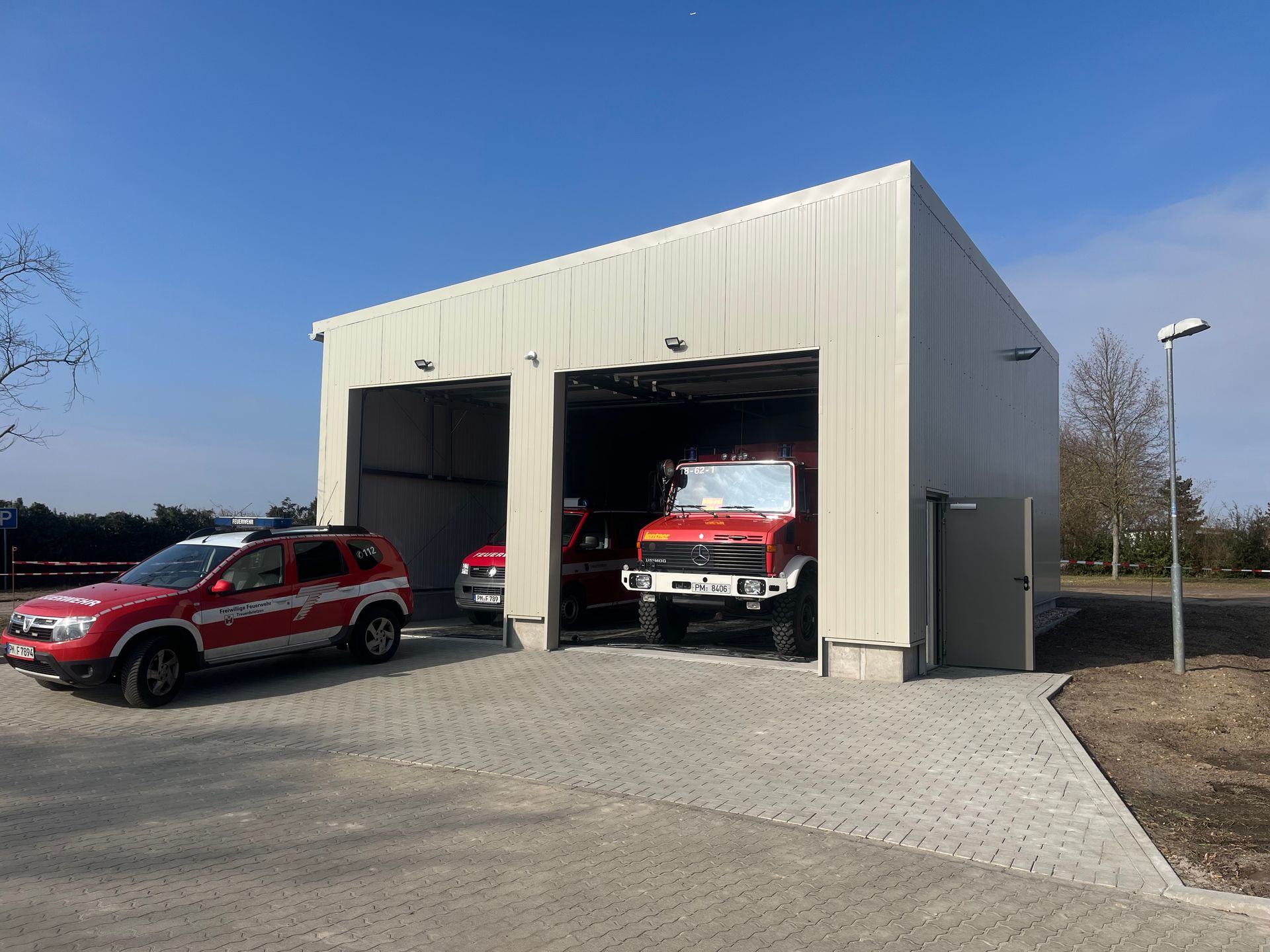 Ein rotes Feuerwehrauto und ein Geländewagen parken an einem sonnigen Tag vor einer beigen Feuerwache.