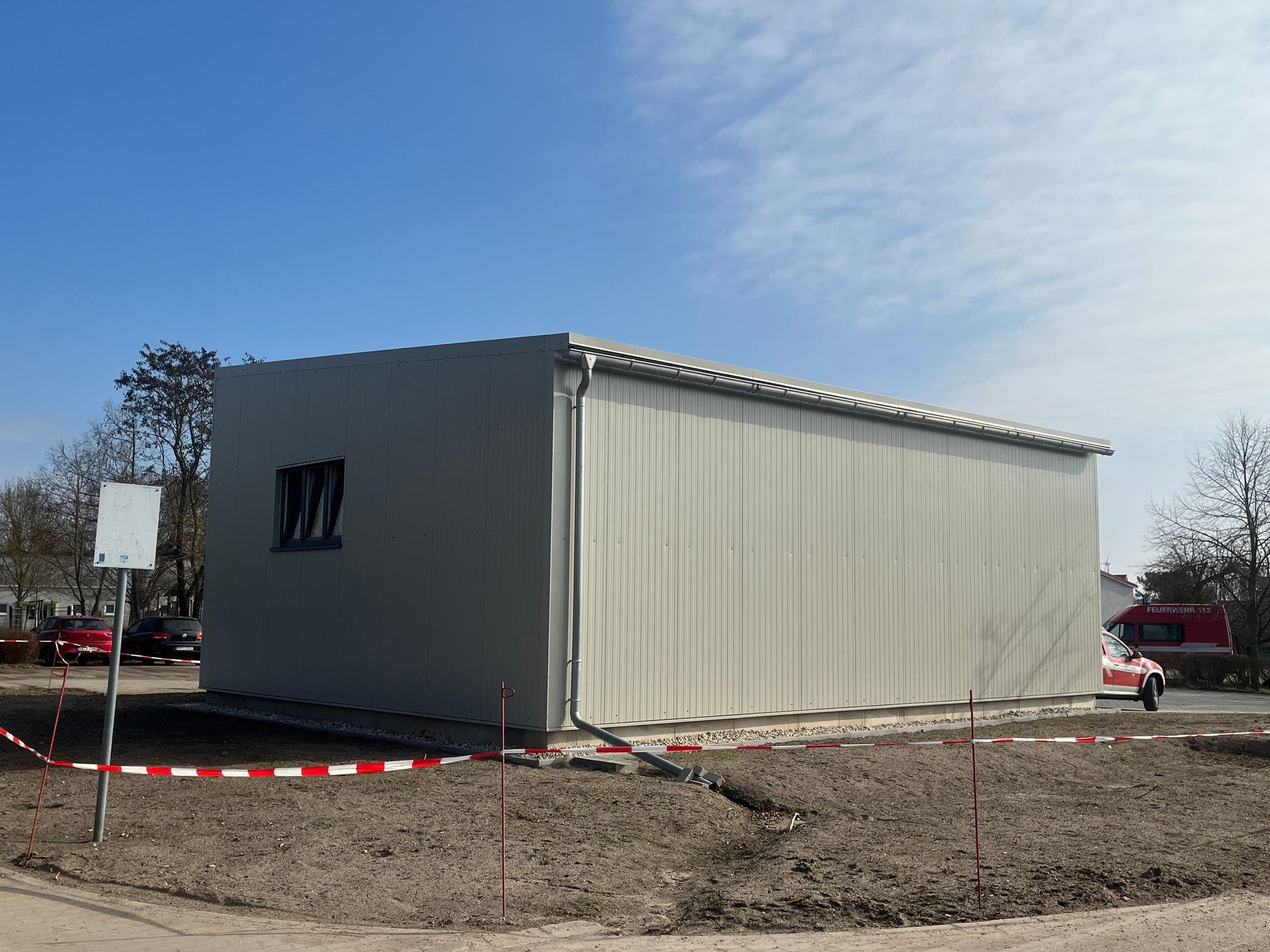 Tragbares graues Modulgebäude auf einem unbefestigten Grundstück, mit rot-weißem Absperrband unter blauem Himmel