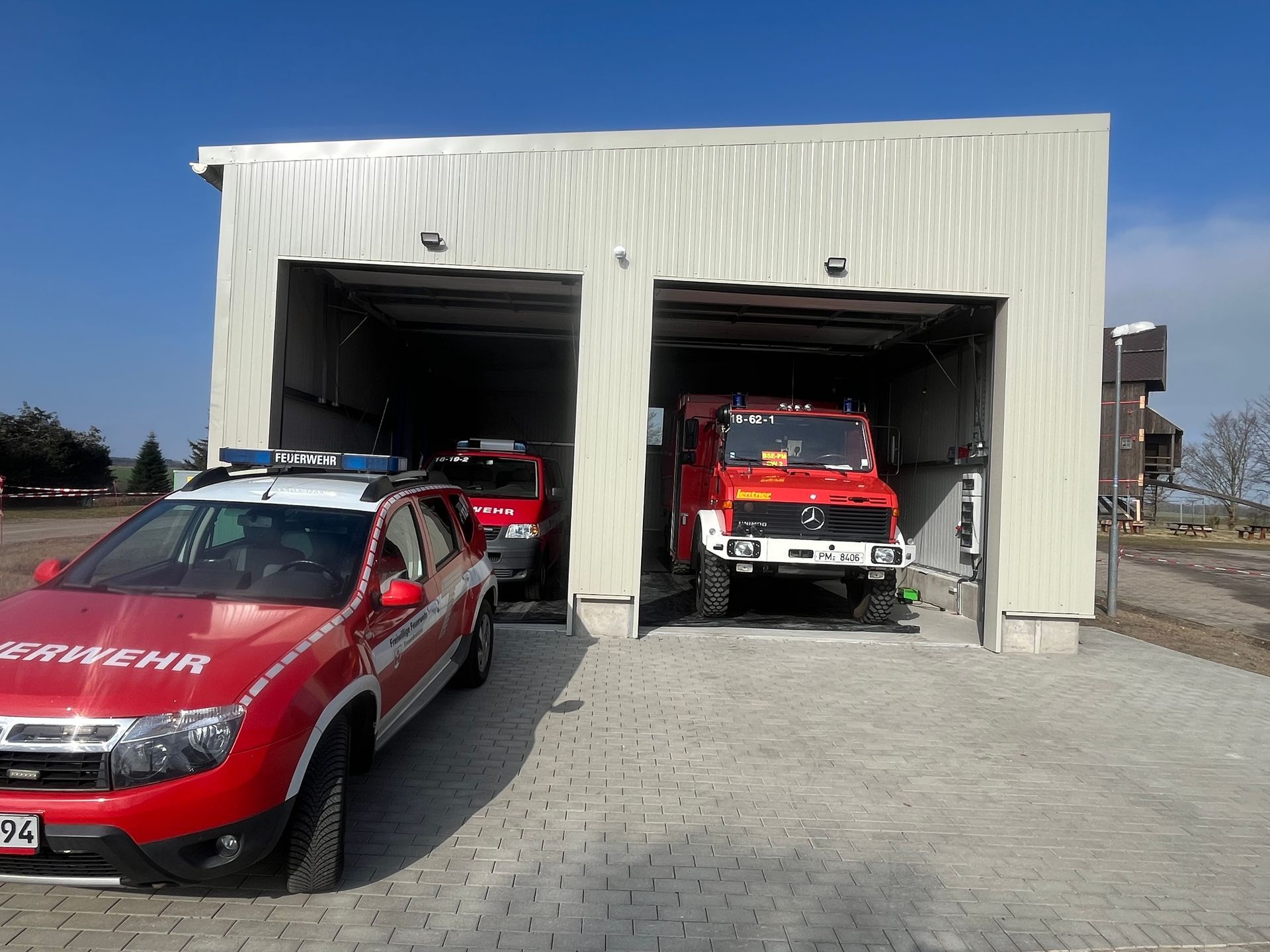 Rote Feuerwehrfahrzeuge parken an einem sonnigen Tag in der Garage einer weißen Feuerwache.