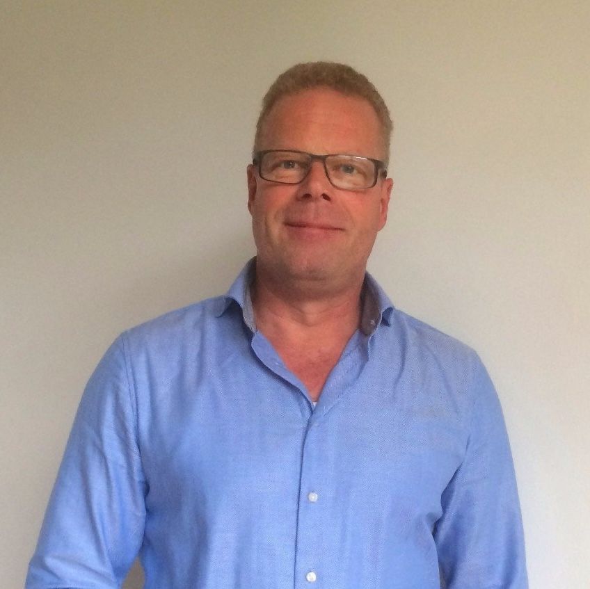 Ein Mann mit Brille und blauem Hemd steht vor einer weißen Wand.