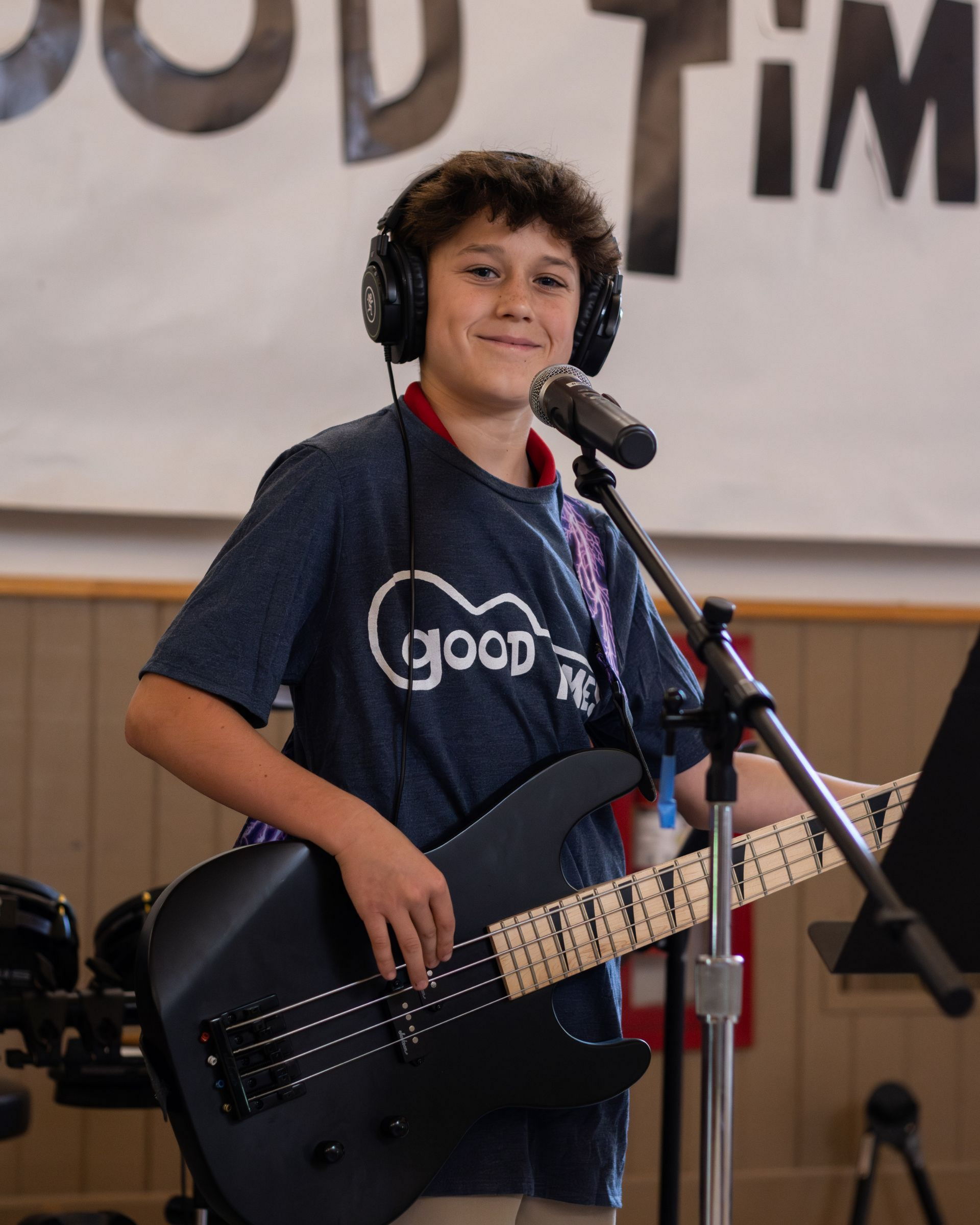 Boy with headphones, holding a bass guitar, singing into a microphone, in front of a banner that says “GOOD TIMES”.