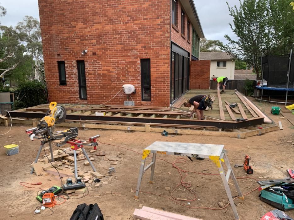 A Man Is Working On A Wooden Deck In Front Of A Brick House — Renovo Contractors In Monash, ACT