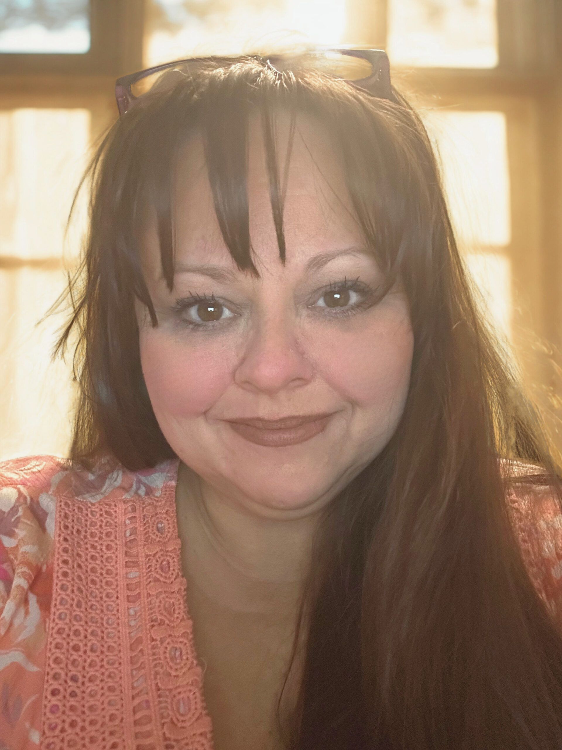 Smiling woman with long dark hair in a pink top, backlit by a window