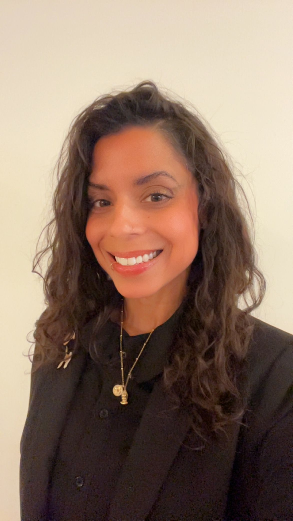 Smiling person with curly hair in a black top against a light wall