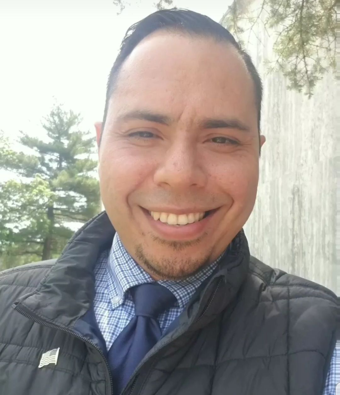 Smiling person outdoors in a black jacket, blue checkered shirt, and navy tie.