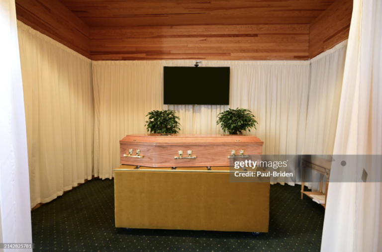 Coffin surrounded by dried grasses, white candles, and white flowers against a green paneled wall