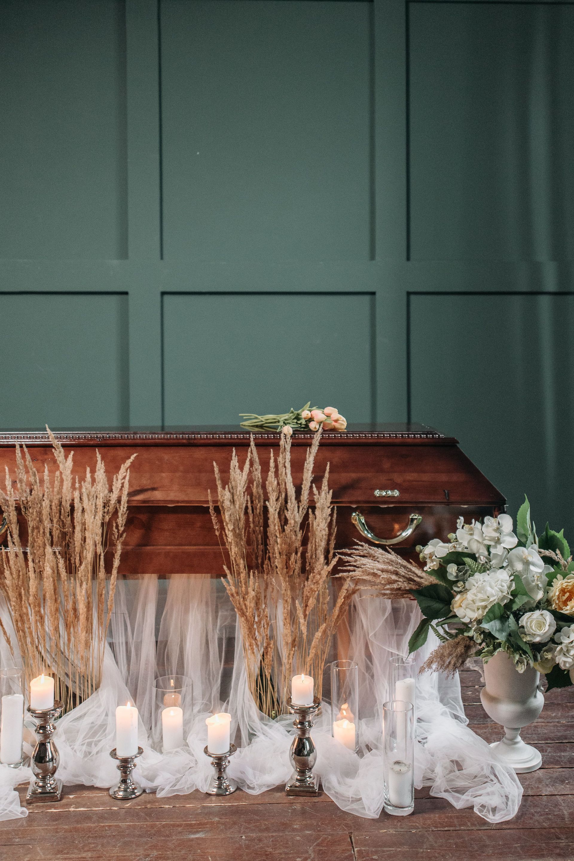 Coffin surrounded by dried grasses, white candles, and white flowers against a green paneled wall
