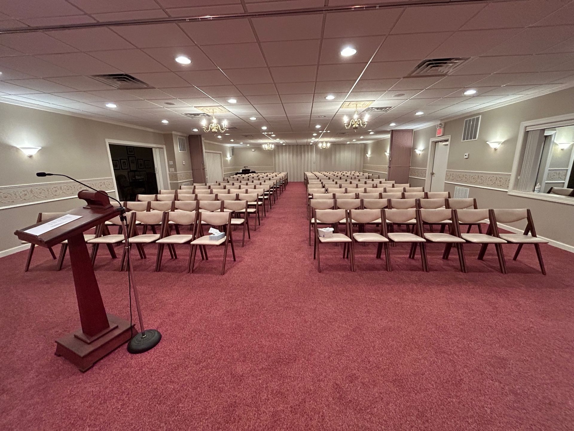 Empty rows of chairs face a wooden podium on red carpet in a well-lit, neutral-toned room.