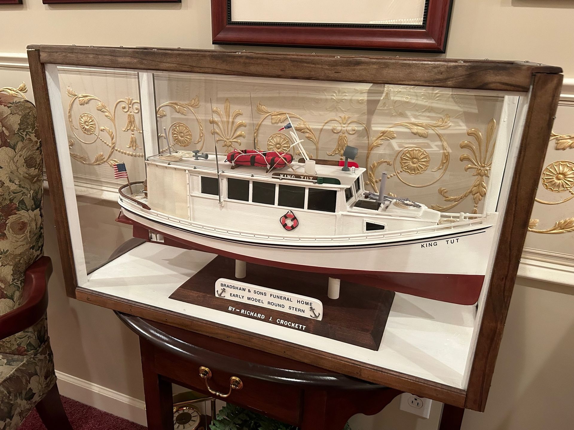 Model boat in a display case on a table. White and red boat, dark wooden frame, and gold wallpaper backdrop.
