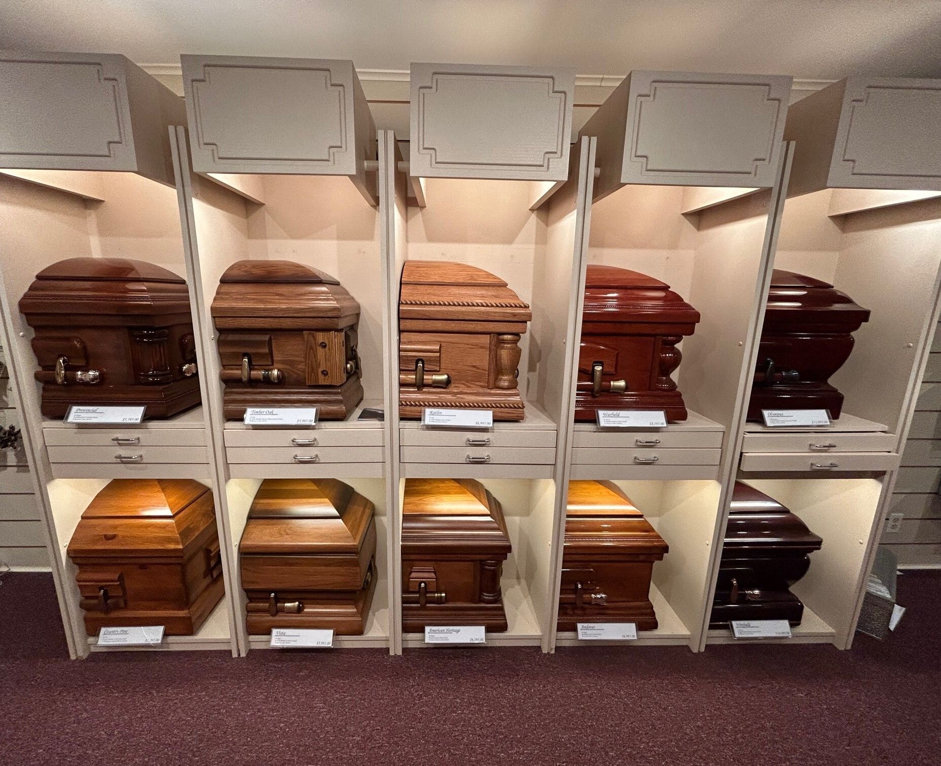 Caskets displayed in a funeral home showroom; various wood tones, lit shelves, and carpeted floor.