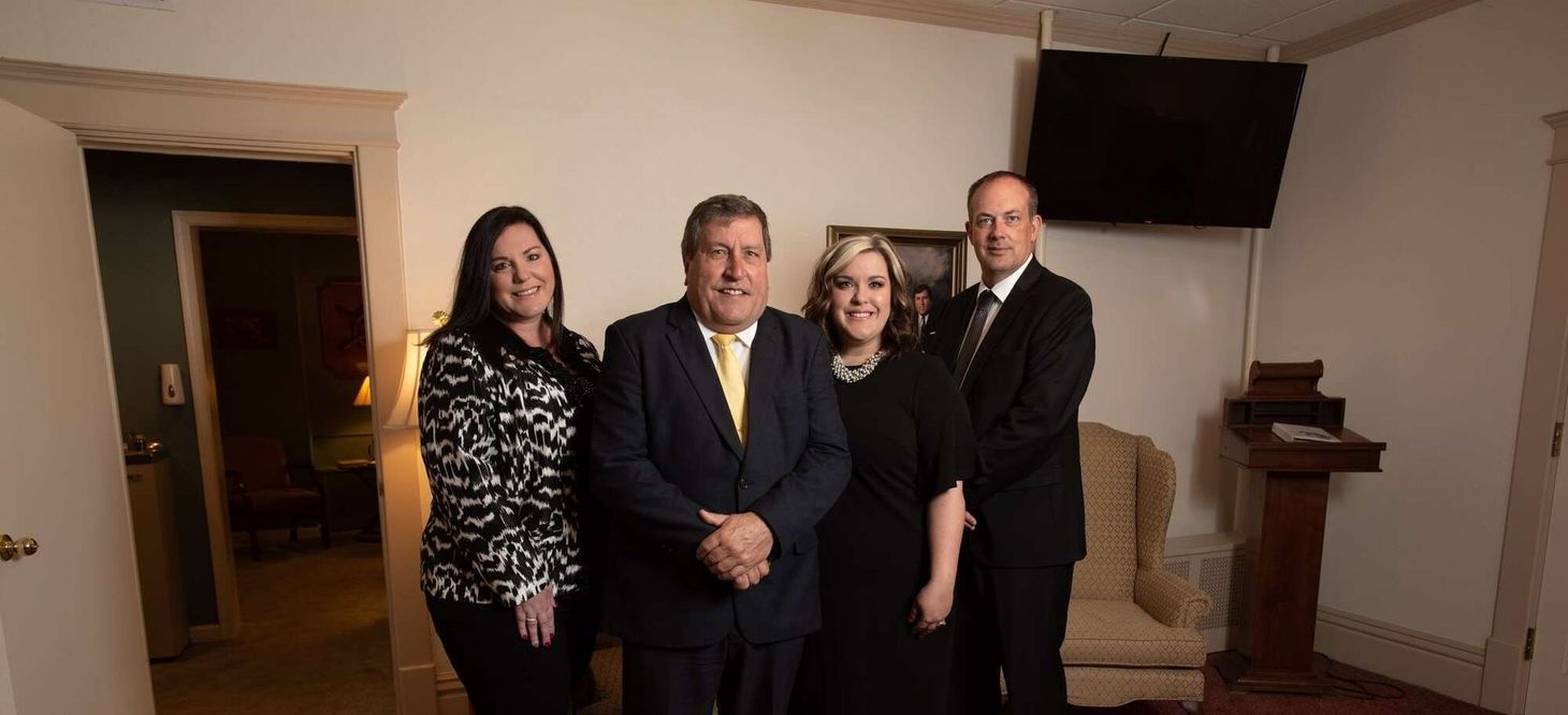 Four people standing in a room, two women, and two men. One man in a navy suit with a yellow tie, the others in black.