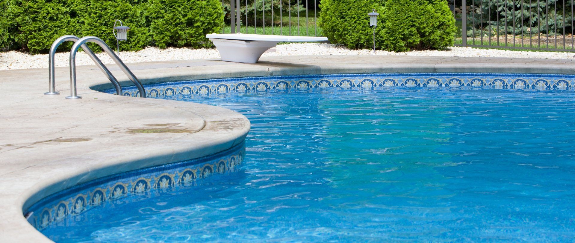 Blue swimming pool with curved edge, metal handrails, and white deck beside green shrubs