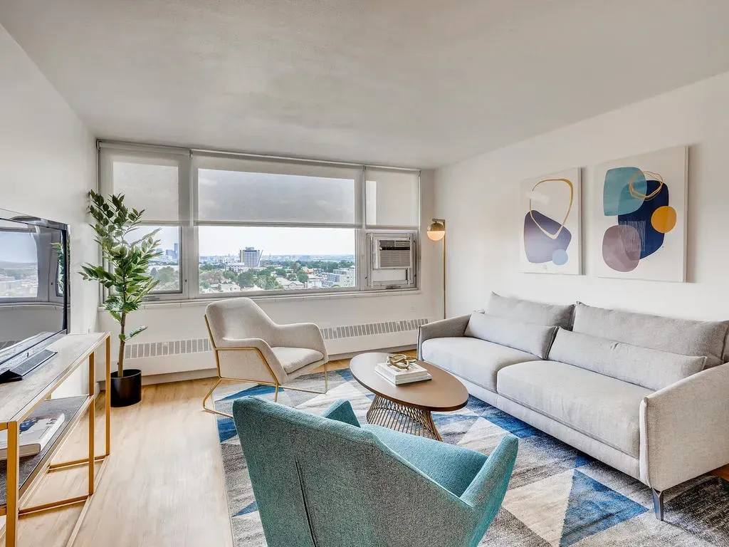 Bright modern living room with large windows, white sofa, teal chair, and city view.