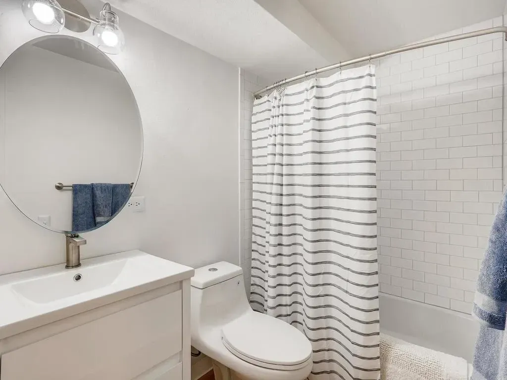 Small white bathroom with vanity, toilet, shower curtain, and round mirror.