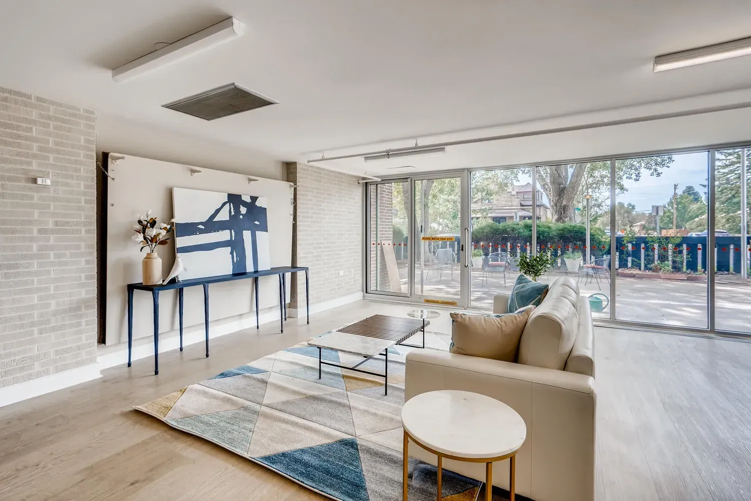Bright modern living room with beige sofa, glass coffee table, wall art, and large windows overlooking a patio.