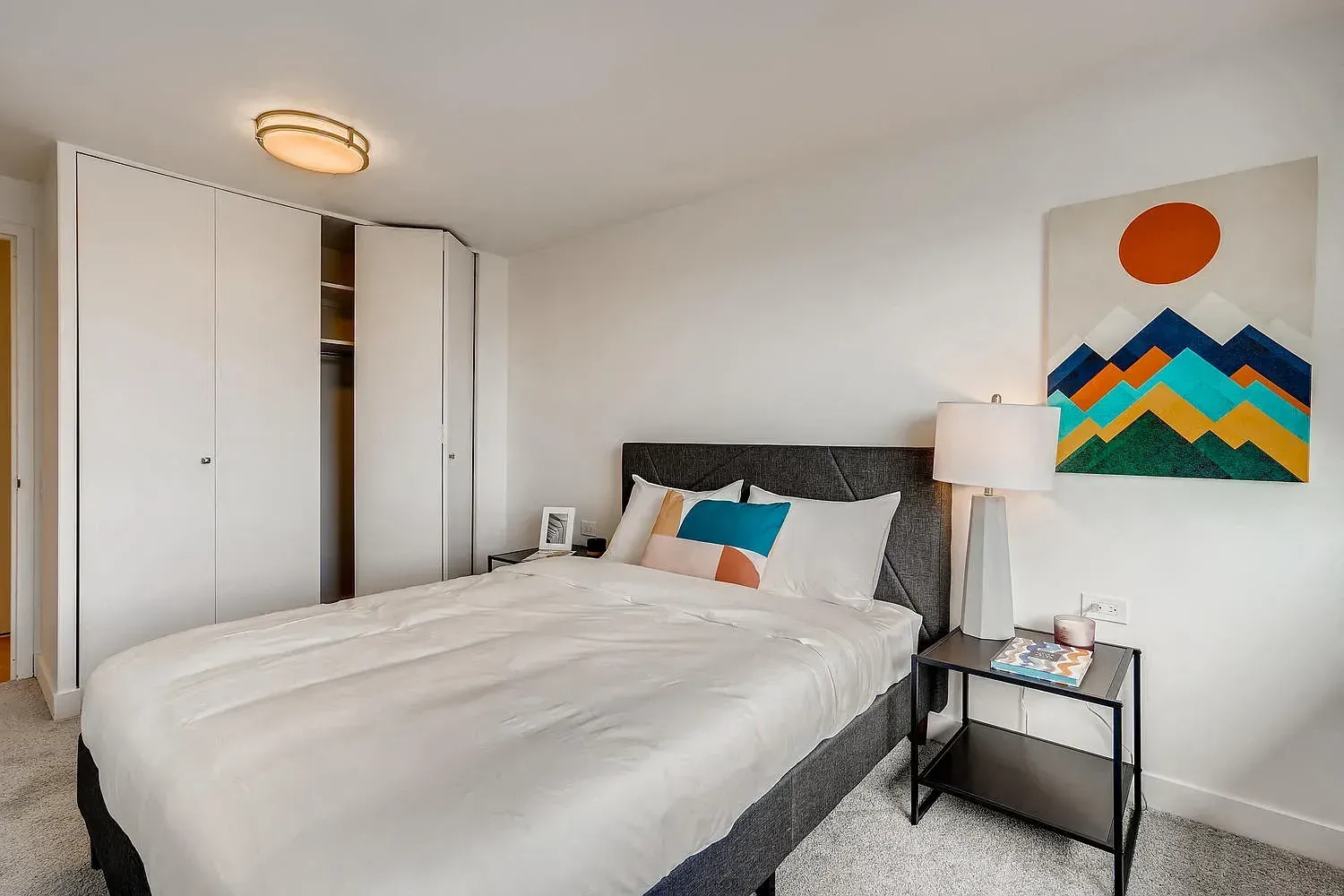 Bright bedroom with a made bed, white closet, and colorful wall art beside a nightstand lamp