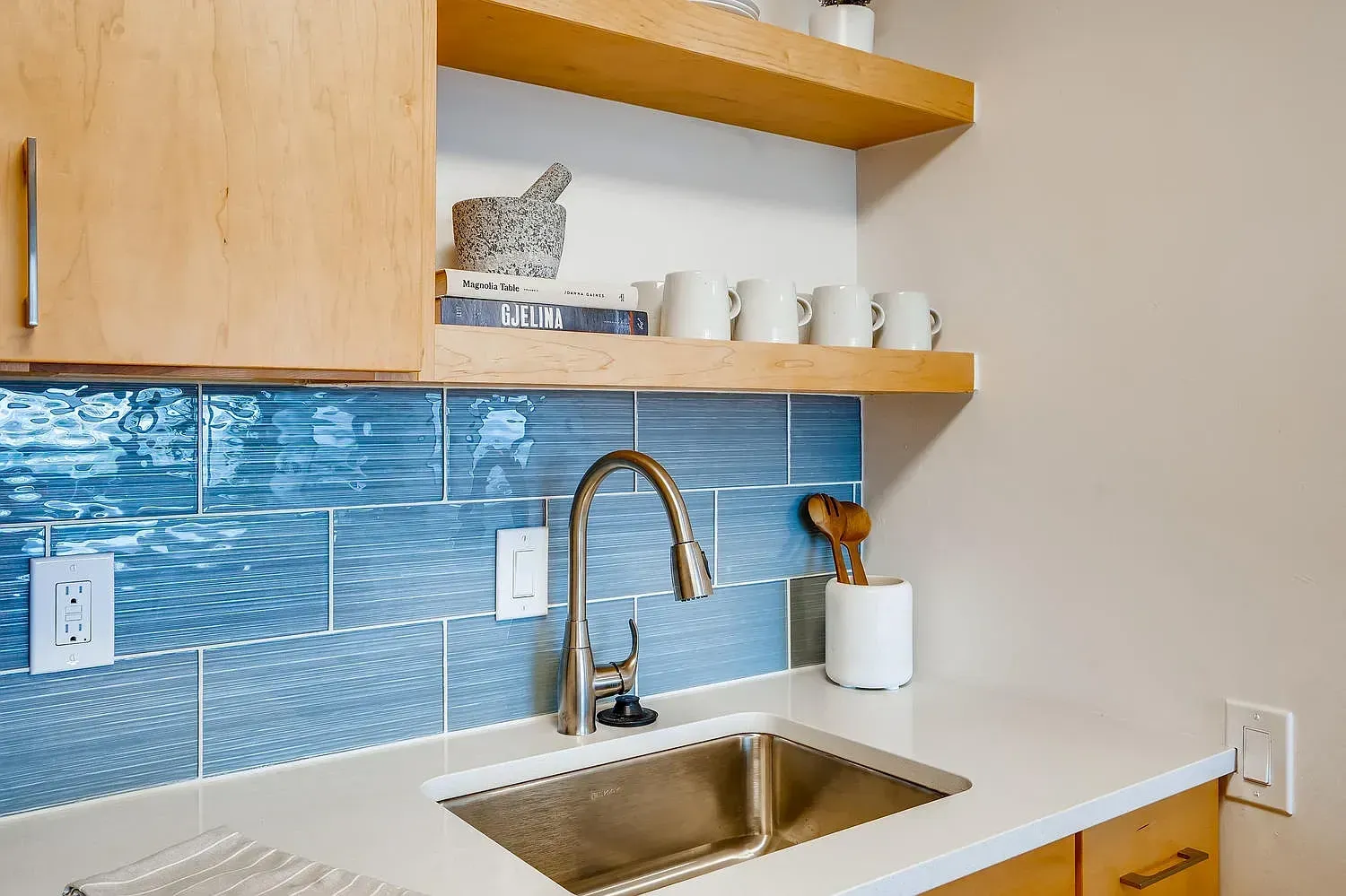 Modern kitchen sink with blue tile backsplash, white cabinets, and wooden shelves