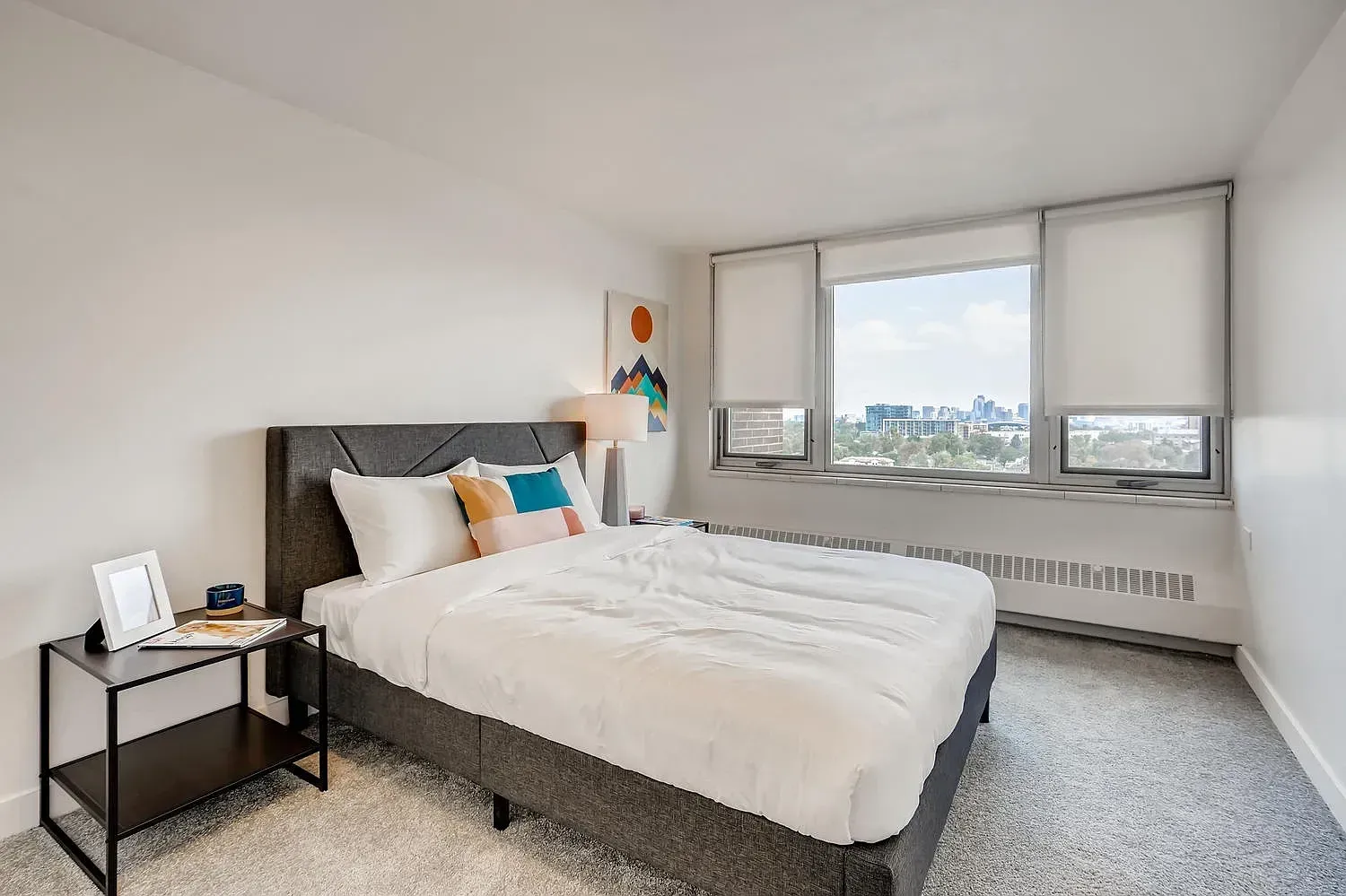 Bright bedroom with a neatly made bed, bedside table, and large window overlooking buildings
