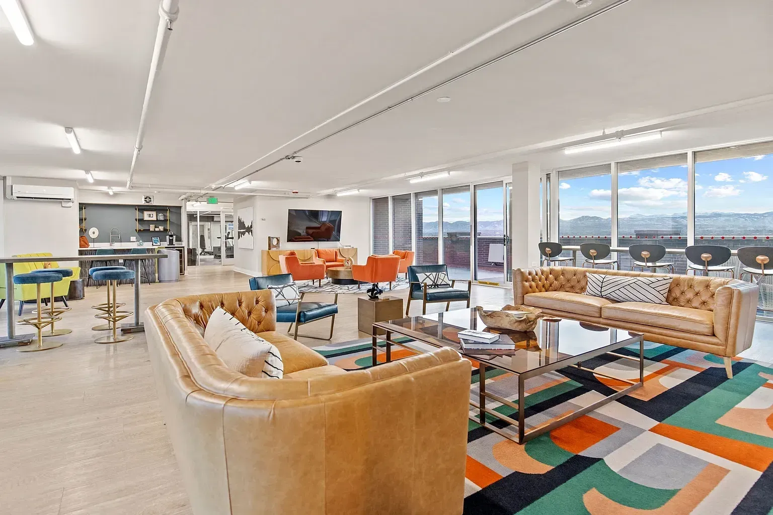 Modern office lounge with tan sofas, colorful rug, high tables, and city-view windows