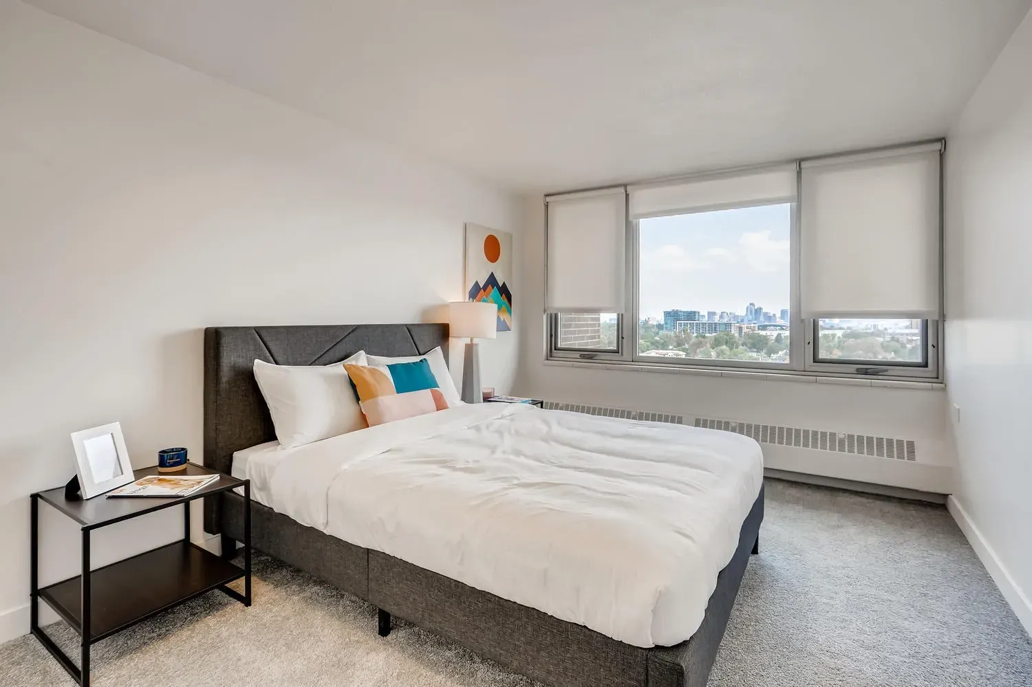 Bright modern bedroom with a white bed, bedside table, and large window overlooking the city.