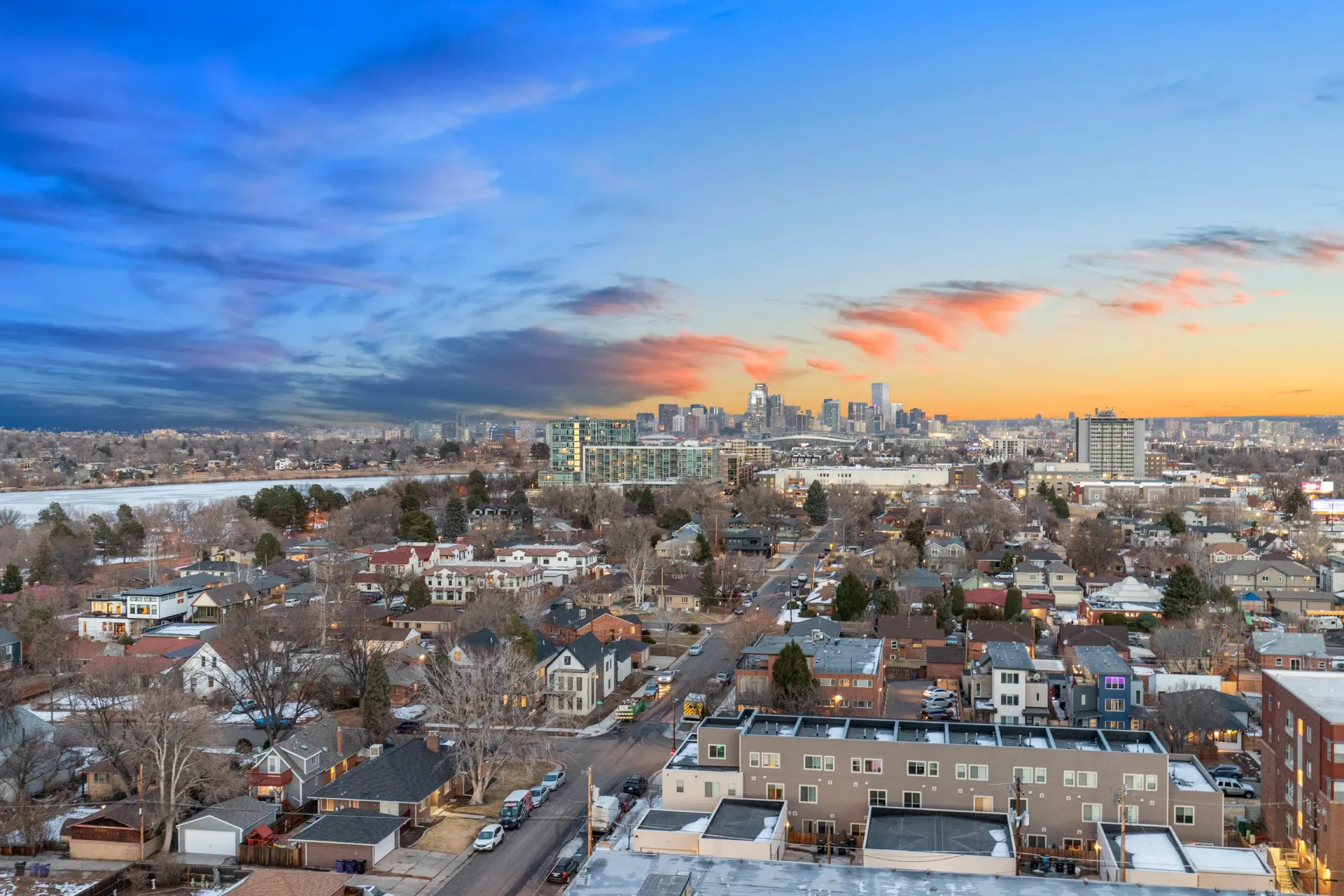 Aerial view of a city skyline at sunset with a lake and residential neighborhood in the foreground.