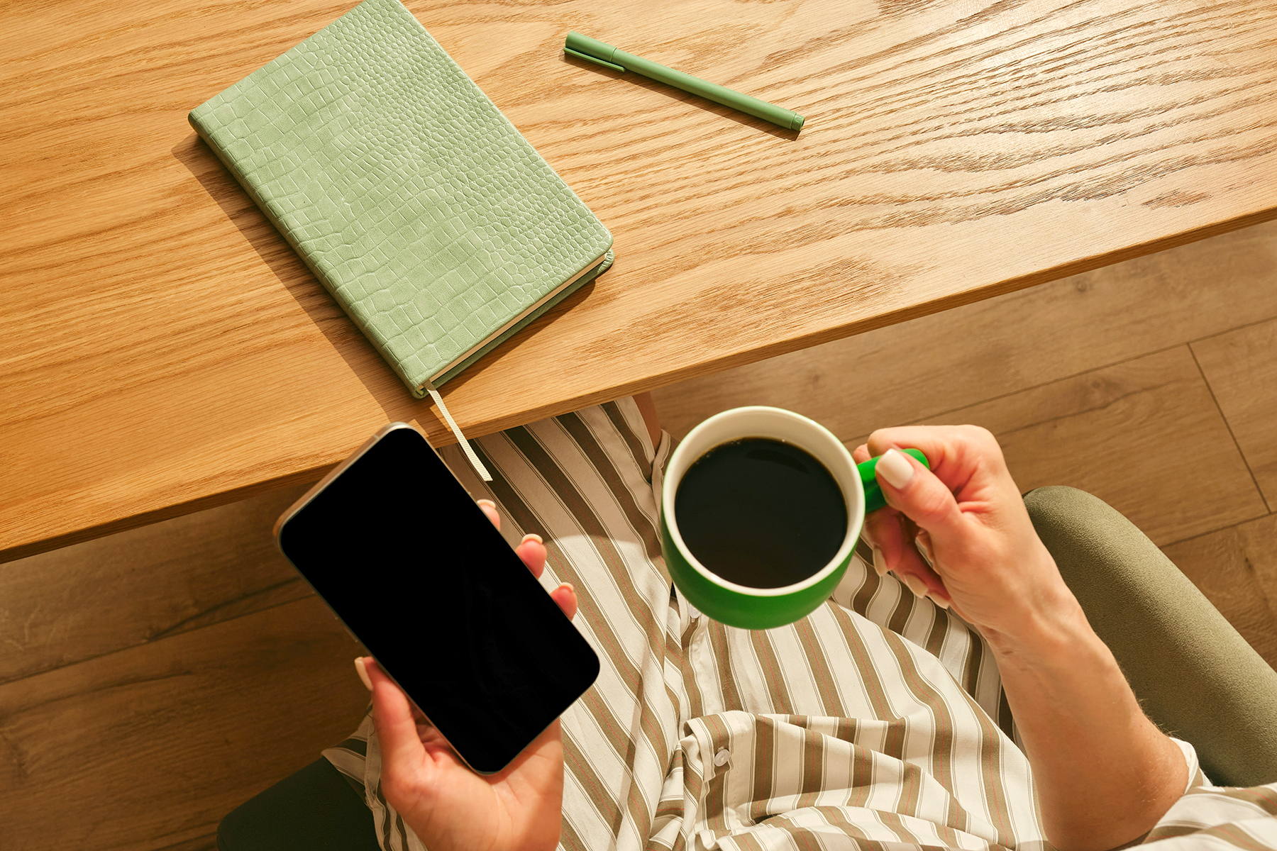 Overhead view of hands holding a phone and coffee beside a notebook and pencil on a wooden desk