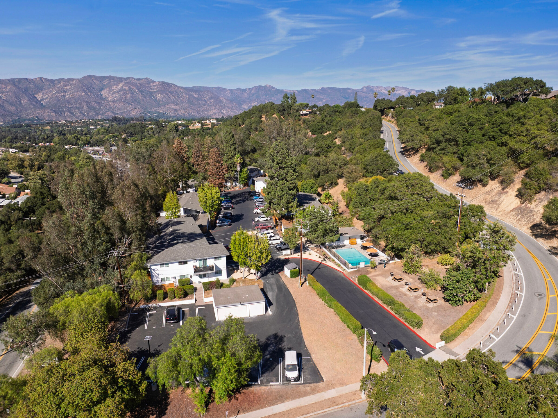 Aerial view of a hillside neighborhood with homes, trees, winding roads, and distant mountains.