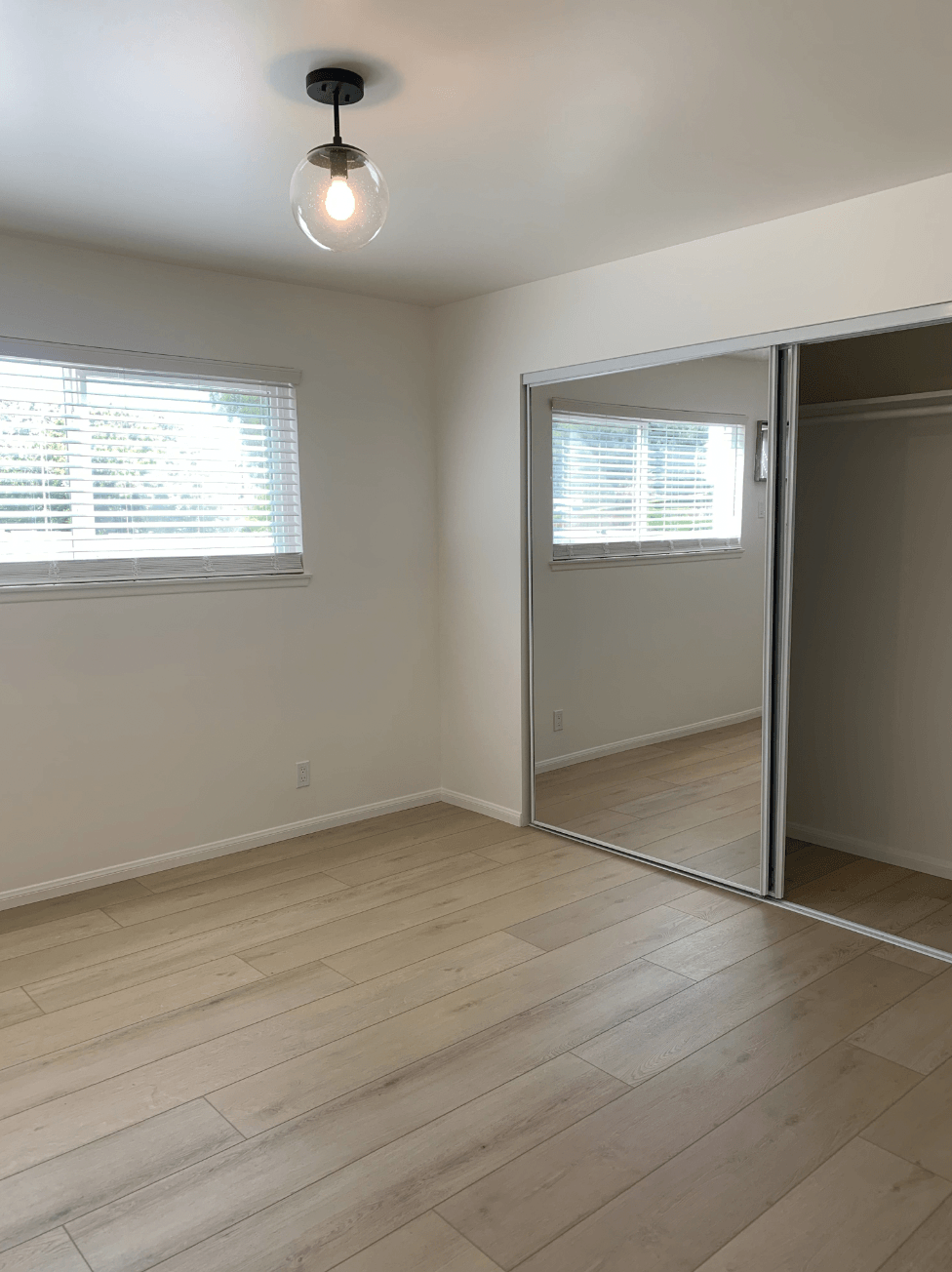 Empty light-filled bedroom with white walls, wood floors, window blinds, and a mirrored closet.
