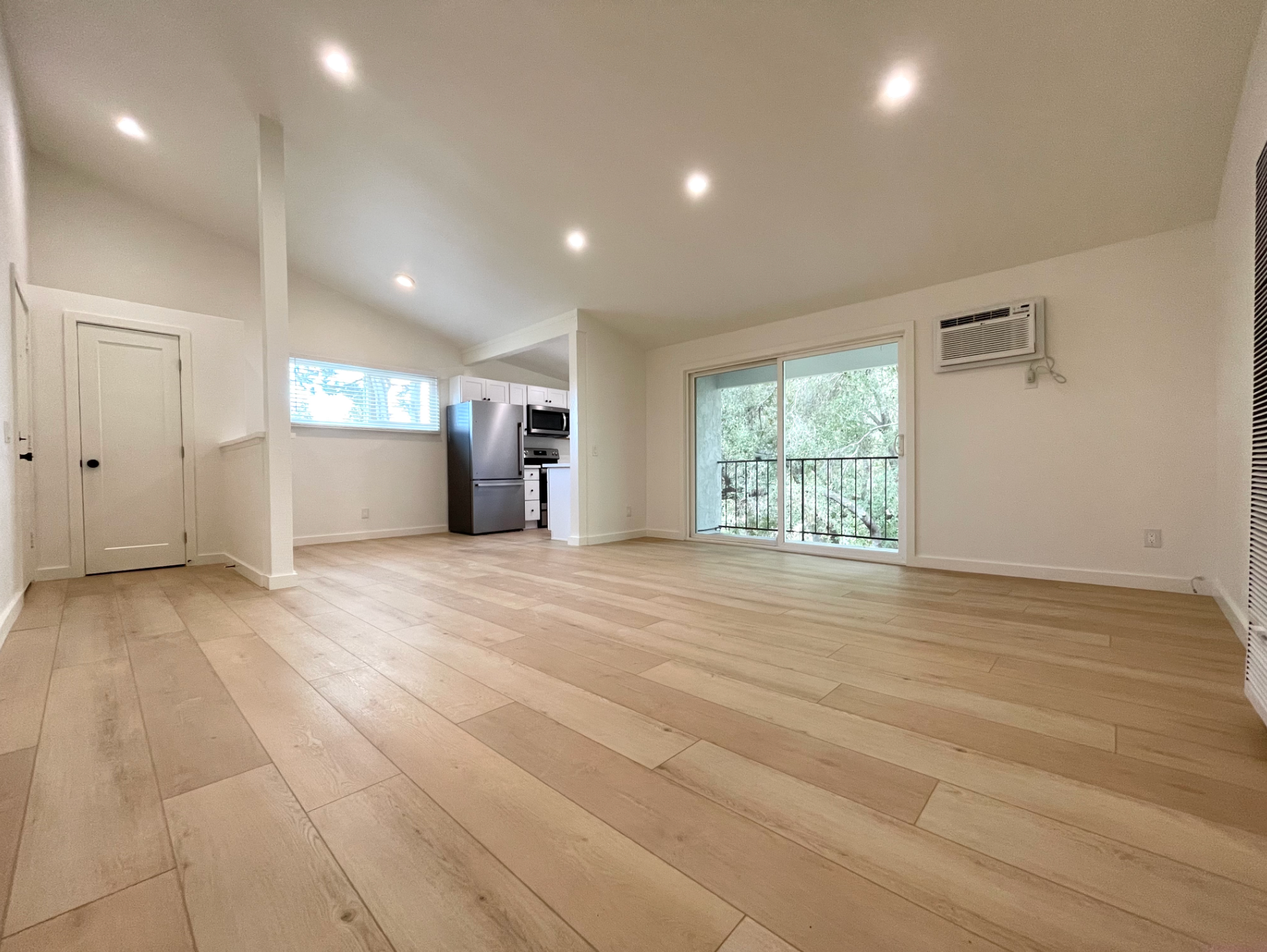 Empty modern living room with light wood floors, white walls, sliding glass door, and recessed lighting
