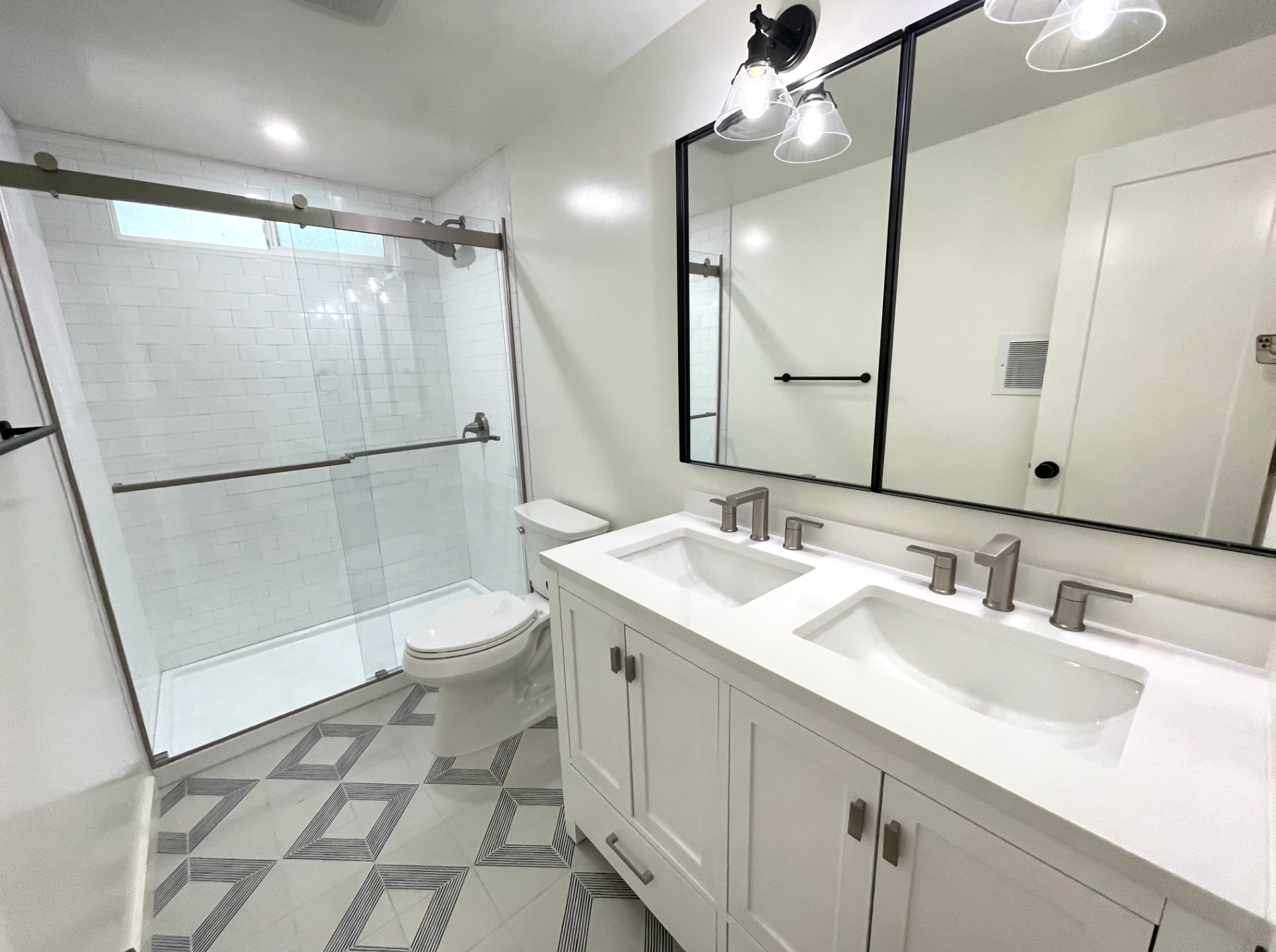 Bright white bathroom with double vanity, large mirror, glass shower, and patterned gray floor tile.