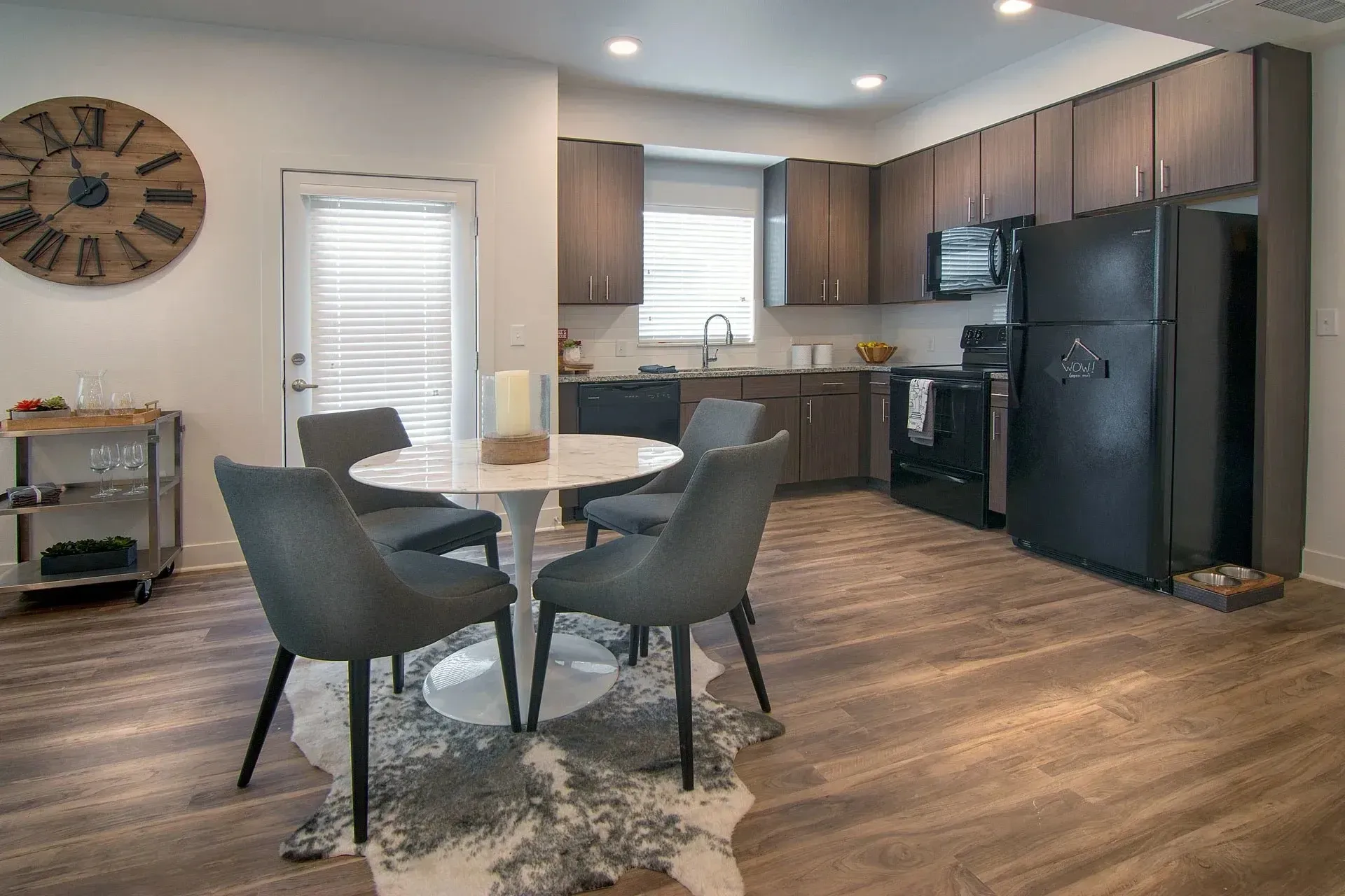Modern dining area and kitchen with dark cabinets, black appliances, and a large wall clock.