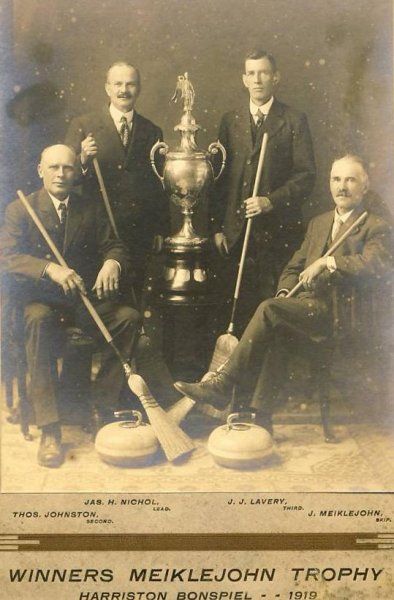 A black and white photo of four men holding curling sticks and a trophy