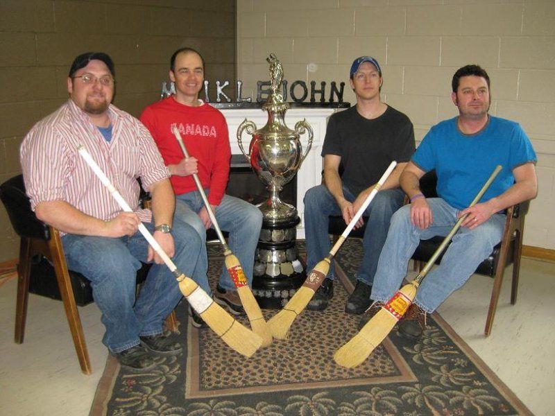 Four men are sitting in front of a trophy and holding hockey sticks
