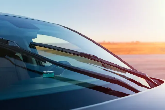 El parabrisas de un coche con los limpiaparabrisas reflejado en una carretera desértica iluminada por el sol al atardecer.